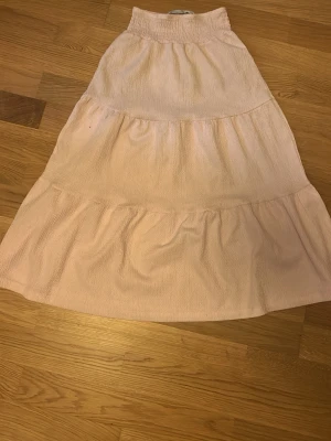 Ljusrosa långkjol i bomull - Säljer en ljusrosa långkjol med volangdetaljer. Kjolen har en mjuk och luftig känsla, perfekt för varmare dagar. Tillverkad i bomull och har en elastisk midja för skön passform. Enkel att matcha med olika toppar för en avslappnad look.