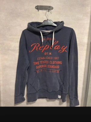 Blå hoodie från Replay - Mörkblå hoodie från Replay med stor röd logga och text på bröstet. Klassisk känguruficka framtill, vita dragsnören i huvan och ribbade muddar vid ärmar och nederkant. Tillverkad i mjukt bomullsmaterial, perfekt för en avslappnad stil. Storlek M som passar 170-180