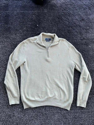 Ralph Lauren half zip - Snygg beige stickad tröja från Polo Ralph Lauren med halv dragkedja och liten broderad logga på bröstet. Tröjan har ribbade muddar vid ärmslut och nederkant samt en hög krage som ger en clean look. Perfekt för lager-på-lager-stil.