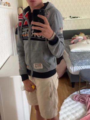 Grå geographical norway hoodie - Säljer en grå Geographical Norway hoodie med dragkedja och flera färgglada tryck och patchar på bröstet och ärmarna. Svarta muddar vid ärmslut och nederkant samt fickor framtill. Perfekt för dig som gillar streetwear och detaljerad design. Är 185 vikt ≈ 68kg. Hör gärna av dig!