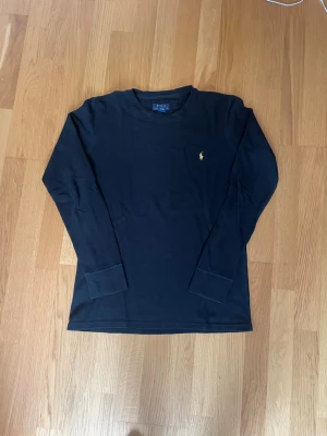 Svart långärmad tröja Polo Ralph Lauren - Klassisk svart långärmad tröja från Polo Ralph Lauren med gul broderad logga på bröstet. Tröjan har rund halsringning och ribbade muddar vid ärmsluten. Perfekt för en clean och stilren look. Den är lite utsliten i kragen