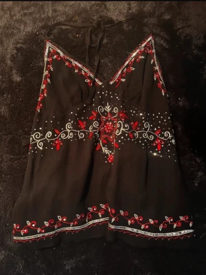 Svart broderad linnetopp med knyt - Svart linnetopp med tunna axelband och knyt i nacken. Toppen har broderade blommor i rött och vitt samt dekorativa detaljer längs kanterna. Perfekt för sommaren och ger en bohemisk känsla. Knyt på ryggen me! Står storlek L men passar snarare s