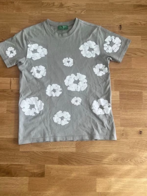 Grå Denim tears t shirt - Säljer en grå t-shirt med vita blommiga tryck över hela plagget. T-shirten har rund halsringning och korta ärmar. Perfekt för dig som vill ha en unik och stilren look med en twist.