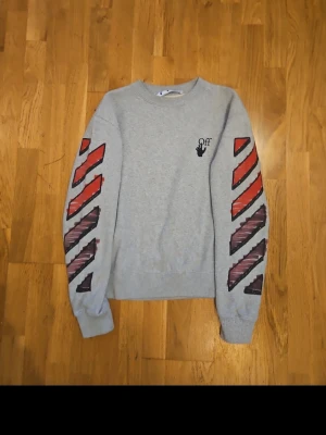 Grå sweatshirt med grafiskt tryck - Säljer en grå sweatshirt med grafiska röda och svarta detaljer på ärmarna och stort tryck på ryggen. Tröjan har rund hals och lång ärm, perfekt för en streetwear-look. Materialet känns som bomull och passformen är normal.