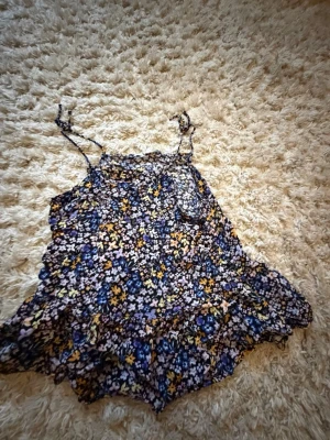 Blommig linnetopp med volang - Söt linnetopp med blommigt mönster i blått, gult och vitt. Toppen har tunna axelband och volangkant nertill, perfekt för en somrig look. Lätt och luftig känsla.