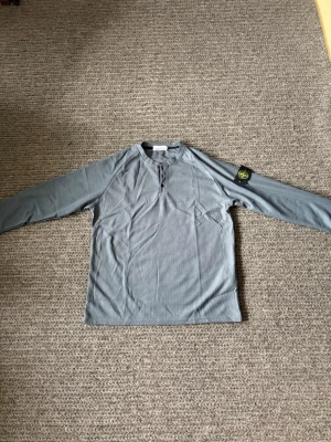Grå långärmad tröja Stone Island - Passar L. Snygg grå långärmad tröja från Stone Island med knappdetaljer vid halsen och den klassiska logotypen på vänster ärm. Tillverkad i ett mjukt och bekvämt material, perfekt för en avslappnad stil. Passar dig som gillar streetwear och enkel design.