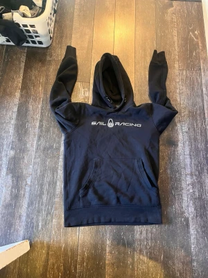 Svart Sail Racing hoodie med tryck - Säljer en svart hoodie från Sail Racing med vitt tryck på bröstet. Klassisk känguruficka framtill och justerbar huva med snören. Mjuk bomullskänsla och ribbade muddar vid ärmslut och nederkant. Perfekt för chill dagar eller när du vill ha en clean look.