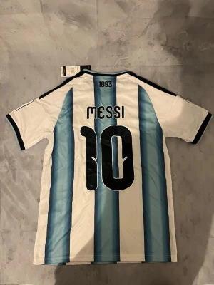 Argentina Messi matchtröja nr 10 - Säljer en officiell Argentina fotbollströja från Adidas med Messi och nummer 10 på ryggen. Tröjan har klassiska blå och vita ränder, svarta detaljer och broderade märken på bröstet. Perfekt för dig som älskar fotboll och vill stötta Argentina.