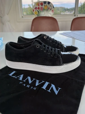 Svarta Lanvin sneakers med lackdetalj - Säljer ett par svarta Lanvin sneakers i mocka med snygga lackade tåpartier och vita, tjocka sulor. Skorna har svarta skosnören och metallfärgade öljetter. Perfekt för dig som gillar stilrena och exklusiva sneakers med en twist. Finns några defekter på bild 2 som man ser på skon de glansiga. Storleken är 39,5 men passar 40.
