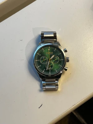 Grön BOSS herrklocka i stål - Snygg herrklocka från BOSS med grön urtavla och silverfärgat armband i rostfritt stål. Klockan har tre mindre urtavlor, datumvisning och tydliga visare. Perfekt accessoar för dig som vill sticka ut med stilren design.