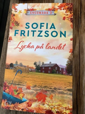 Lycka på landet av Sofia Fritzson - En charmig och idyllisk roman av Sofia Fritzson, tredje delen i serien om byn Ängsmark. Omslaget har höstfärger med gula och orange löv, en lantlig gård och ett blått paraply. Perfekt för dig som gillar romantik och småstadsliv.