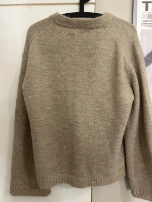 Beige stickad tröja med rund hals - Mysig beige stickad tröja med rund halsringning och långa ärmar. Tröjan har en enkel, rak passform och ribbstickade muddar vid ärmslut och nederkant. Perfekt för lager på lager under kyliga dagar.