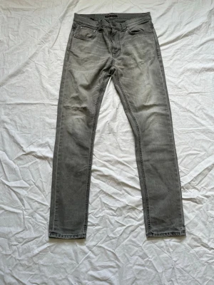 Grå jeans från Nudie Jeans Co - Säljer ett par grå jeans från Nudie Jeans Co i modellen Lean Dean. Jeansen har en slim fit, klassisk femficksdesign och ett litet slitmärke på vänster lår. Tillverkade i mjuk denim med snygg tvättad look.