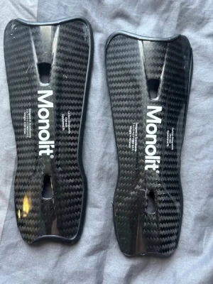 Monolit benskydd i kolfiber - Säljer ett par svarta benskydd från Monolit i kolfiber med snyggt mönster och vit logga. Lätta och smidiga med ergonomisk form som passar perfekt för fotboll eller annan sport där skydd behövs.
