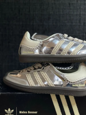 Adidas Wales Bonner silvriga sneakers - Unika sneakers från Adidas x Wales Bonner med glansig silverfärgad ovandel, krämvita detaljer och stickade ränder. Låg modell med mörk sula och Wales Bonner-logga i guld på sidan. Kombination av syntetmaterial och textil ger en modern och trendig look.