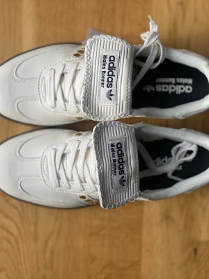 Adidas Bälte Bouncer vita sneakers - Snygga vita Adidas Bälte Bouncer sneakers med klassisk design och bred plös. Skorna har vita snören, detaljer i guld och svart logga på plösen. Ovandelen är i läder och textil, och sulan är i gummi. Perfekta för dig som gillar retrostil och streetwear.