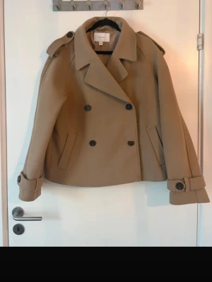 Trendig beige/brun kort kappa🍂 - Stilren och höstig beige kort kappa från VILA med dubbelknäppning, bred krage och axelklaffar. Rocken har två snedställda fickor och dekorativa spännen vid ärmsluten. Tillverkad i ett mjukt material som ger en klassisk look. Köpt på Vinted men aldrig Andvänd av mig. Bilderna är ifrån förra ägaren.  Lite nopprig men det går säkert att få bort enkelt med en noppkamm. Annars i fint skick. Kan alltid diskutera priset så skriv bara!