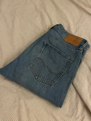 Relaxed/Chris jeans från Jack & Jones  - Klassiska blå jeans från Jack & Jones i rak modell. Jeansen har en ljus tvätt, fem fickor och detaljer i kopparfärgad metall. Tillverkade i slitstark denim med en snygg läderpatch bak i midjan.