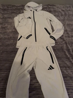 Vit Adidas träningsdress med huva - Säljer en vit träningsdress från Adidas med svarta detaljer. Setet består av en jacka med huva, dragkedja och två fickor samt matchande byxor med resår i midjan och dragkedjeförsedda fickor. Perfekt för träning eller chill. Materialet känns lätt och sportigt.