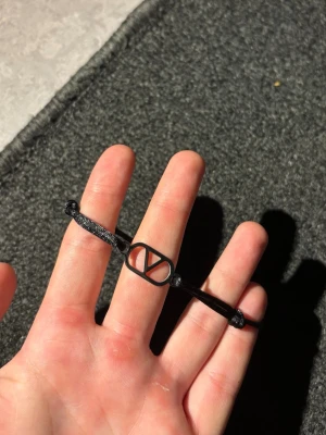 Valentino armband - Snyggt svart armband med justerbart band och en metallogo i mitten. Logotypen har en stilren design och armbandet är gjort av textil och metall. Perfekt accessoar för dig som gillar minimalistisk och modern stil.