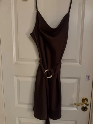 Brun omlottklänning med bälte - Snygg brun kortklänning med tunna axelband och omlottdesign. Klänningen har ett matchande tygbälte med rund spänne i midjan och ett mjukt, glansigt material som ger en elegant look.