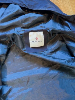 Svart dunjacka från Moncler - Säljer en mörkblå dunjacka från Moncler i storlek L. Jackan har klassisk Moncler-logga på insidan och ett unikt serietidningsmärke. Tillverkad i glansigt material som ger en exklusiv känsla. Perfekt för kalla vinterdagar.