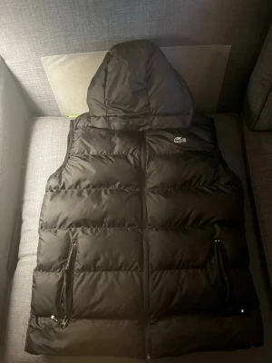 Svart dunväst med huva från Lacoste - Snygg svart dunväst från Lacoste med huva och dragkedjor på fickorna. Västen har en quiltad design och den klassiska Lacoste-loggan på bröstet. Perfekt för lager-på-lager och streetstyle. Fast pris, Medium men passar Large