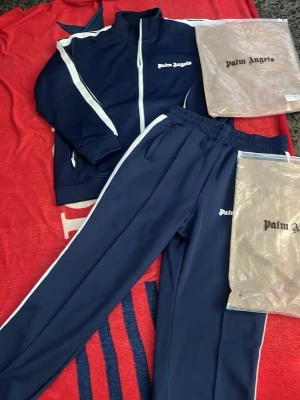 Mörkblåa mjukisbyxor Palm Angels - Säljer ett par mörkblåa mjukisbyxor från Palm Angels med vita sidoränder och logga på benet. Byxorna har resår i midjan och är gjorda i ett mjukt, sportigt material. Perfekta för en chill och trendig look. Pris kan diskuteras 👍👍