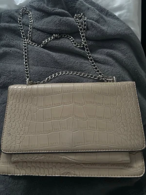 Beige axelremsväska med kedja - Stilren beige handväska i croco-mönstrat konstläder med silverfärgad kedjerem. Väskan har ett lock med magnetknäppning och flera fack inuti för bra organisering. Perfekt accessoar för att lyfta din outfit.