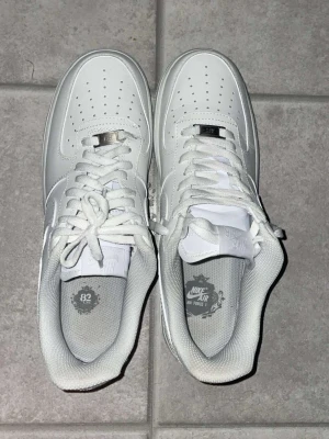 Nike Air Force 1 vita låga sneakers - Klassiska Nike Air Force 1 sneakers i helvitt läder med perforerad tåbox, vita skosnören och diskret Nike-logga på sidan. Skorna har en chunky sula och metallbricka på snörningen. Perfekt för dig som gillar stilrena och tidlösa sneakers.