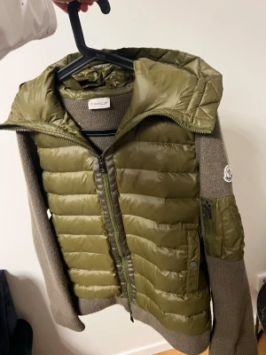 Olivgrön cardiganjacka från Moncler - Snygg olivgrön cardiganjacka från Moncler med quiltad front och huva i glansigt material. Ärmar och rygg i stickad ullmix, dragkedja framtill och fickor med knapp. Moncler-logga på ärmen och extra ficka på armen. Perfekt för lager-på-lager-stil.