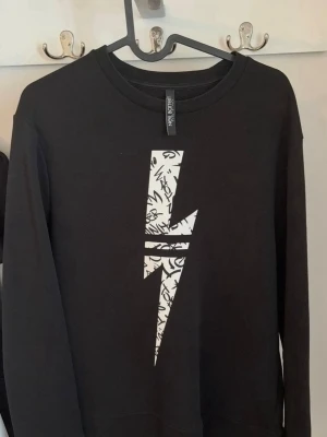 Svart sweatshirt med blixt från Neil Barrett - Cool svart sweatshirt från Neil Barrett med vit blixt på bröstet och grafiskt mönster i trycket. Tröjan har rund hals, långa ärmar och är gjord i mjuk bomullsblandning (65% bomull, 35% polyester). Slim fit-modell. 