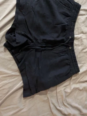 Mörkblå shorts med snörning - Ett par mörkblå shorts med elastisk midja och snörning framtill. Shortsen har en enkel design och är tillverkade i ett mjukt material, perfekta för avslappnade dagar. Passformen är normal och de har fickor på sidorna.