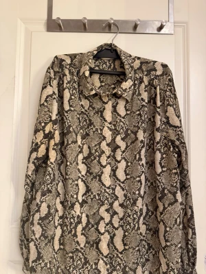 Beige och svart skjorta med ormmönster - Säljer en långärmad skjorta med beige och svart ormmönster. Skjortan har klassisk krage och knappar framtill. Perfekt för dig som vill sticka ut med ett djurmönster i garderoben.