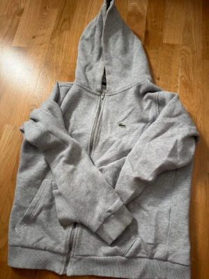 Grå zip hoodie från Lacoste - Säljer en grå zip hoodie från Lacoste med klassisk krokodillogo på bröstet. Hoodien har dragkedja framtill, två fickor och ribbade muddar vid ärmar och nederkant. Perfekt för en avslappnad och stilren look.