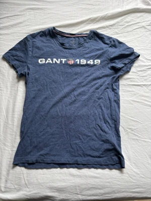 GANT t-shirt  - Säljer en blå t-shirt från GANT med vitt tryck 'GANT 1949' och en liten sköld på bröstet. Klassisk rund hals och korta ärmar. Perfekt till jeans eller shorts för en avslappnad look.