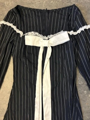 Svart långklänning med vit rosett - Snygg svart miniklänning med vita vertikala ränder, långärmad design och offshoulder-skärning. Klänningen har en stor vit rosett framtill och spetsdetaljer längs axlarna. Perfekt för dig som gillar unika detaljer och klassisk stil.