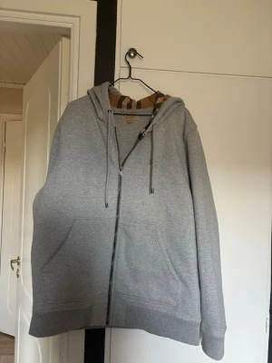 Grå zip hoodie med huva Burberry  xl  - En stilren äkta Burberry grå hoodie med dragkedja framtill och två stora fickor. Tröjan har det klassisk mönstret på huvan.. Perfekt för en avslappnad look och enkel att matcha med det mesta. Du ser att den är äkta på den bruna lappen där storleken visas, jag köpte fel storlek då vart jag än läste så skrev man: ”Burberry kan vara mindre i storlek ” så jag som ville att det skulle bli så bra som möjligt tog en större storlek- det hade inte behövts. Likt Burberrys ”sjuka” så är dragkedjan lagad. 