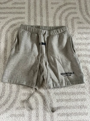 Grå Essentials Shorts - Säljer ett par grå mjukis shorts från Essentials med snörning i midjan och sidofickor. Mjuk bomullskänsla och tryckt logga på ena benet. Perfekta för chill dagar eller träning.