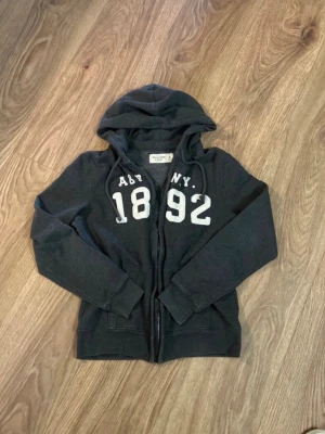 Abercrombie & Fitch hoodie - Snygg mörkgrå hoodie från Abcrombie & Fitch med dragkedja och huva. Tryck i vitt framtill med texten 'ABY NY 1892'. Mjuk insida och två fickor framtill. Perfekt för en avslappnad streetwear-look.
