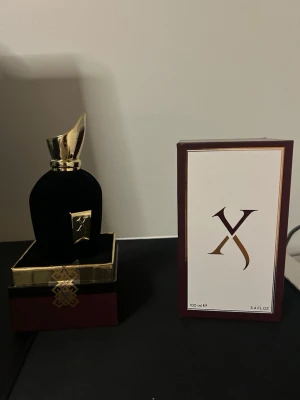 Lyxig svart parfymflaska med gulddetaljer - Exklusiv parfym i en elegant svart flaska med sammetsyta och guldfärgat lock. Flaskan har en unik form med en snedställd topp och en guldfärgad etikett framtill. Kommer i en stilren ask och med matchande förpackning med guld- och vinröda detaljer.