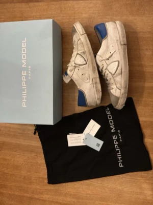 Vita Philippe Model sneakers med blå detaljer i storlek 42 - Snygga vita sneakers från Philippe Model med blå detaljer på häl och plös. Skorna har klassisk låg modell, vita snören och diskret logga på sidan. Tillverkade i läder med mockadetaljer och robust sula..