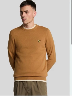 Kamelbrun tröja med broderad logga - Snygg kamelbrun tröja i mjukt material med rund halsringning och ribbade muddar. På bröstet sitter en broderad gul logga som ger en clean och stilren look. Perfekt att matcha med jeans eller chinos för en avslappnad stil.