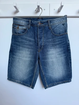 Blå jeansshorts från Solid - Snygga blå jeansshorts från Solid med klassisk femficksdesign och slitna detaljer. Shortsen har normal passform, bälteshällor och knappgylf. Perfekta för en avslappnad stil under varma dagar. Mycket bra skick, nyskick.