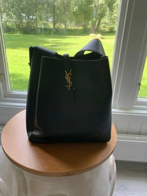 Svart Saint Laurent handväska - Snygg svart handväska från Saint Laurent i slätt läder med guldigt YSL-emblem framtill. Väskan har en stilren bucket-modell och bred axelrem som gör den enkel att bära. Perfekt för dig som gillar klassisk och lyxig design.