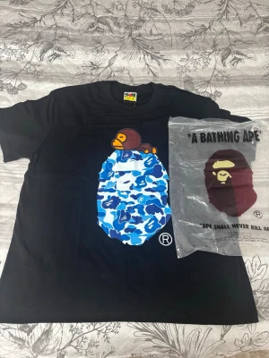 Svart BAPE t-shirt med blått camomönster - Säljer en svart t-shirt från A Bathing Ape med stort tryck av en apa och blått camomönster på bröstet. Klassisk passform och rund hals. Perfekt för dig som gillar streetwear och vill sticka ut med en ikonisk design.
