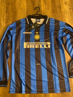 Inter Milan långärmad matchtröja Umbro - Säljer en långärmad Inter Milan matchtröja från Umbro i blått och svart med klassiska vertikala ränder. Tröjan har broderad klubbmärke, gul stjärna, Pirelli-tryck och vit Umbro-logga. Tillverkad i lätt och ventilerande material, perfekt för fotboll eller samling.