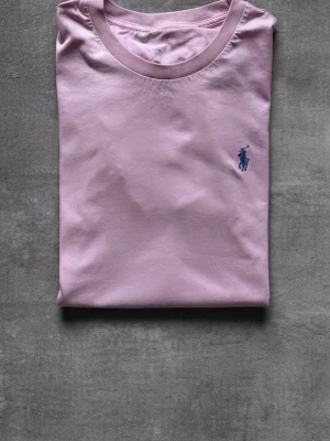 Ljusrosa t-shirt från Polo Ralph Lauren - En stilren ljusrosa t-shirt från Polo Ralph Lauren med klassisk passform och korta ärmar. T-shirten har en liten blå broderad logga på bröstet och är tillverkad i mjuk bomull. Perfekt för en clean och avslappnad look.
