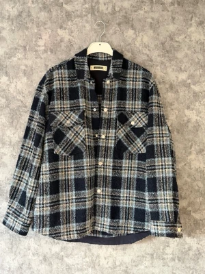 Rutig overshirt från Woodbird - Snygg overshirt i tjockt material från Woodbird. Mörkblå och beige rutigt mönster, två stora bröstfickor med knappar och klassisk krage. Perfekt lager på lager-plagg för en trendig look. Och som passar perfekt nu till hösten! 