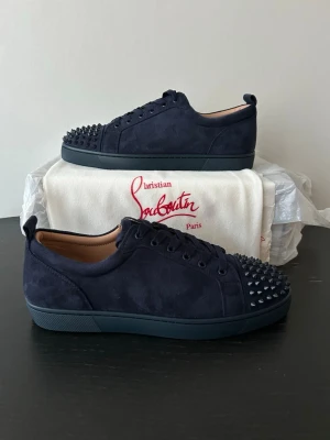 Mörkblå sneakers med nitar Louboutin - Snygga mörkblå sneakers i mocka från Christian Louboutin med svarta nitar på tån och den klassiska röda sulan. Skorna har lågt skaft, matchande mörkblå snörning och en exklusiv känsla. Perfekta för dig som vill sticka ut med stil. BARA OG DUSTBAG MEDFÖLJER 